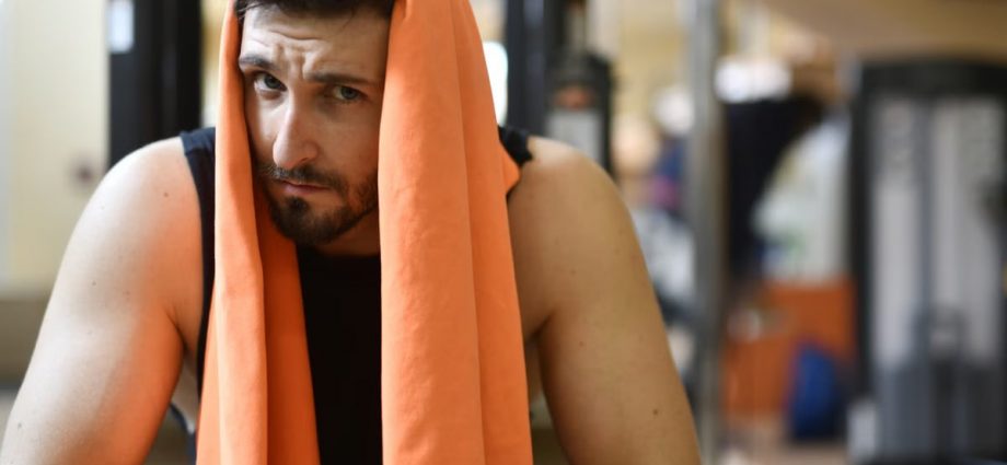 A tired man rests with an orange towel after a workout in the gym.