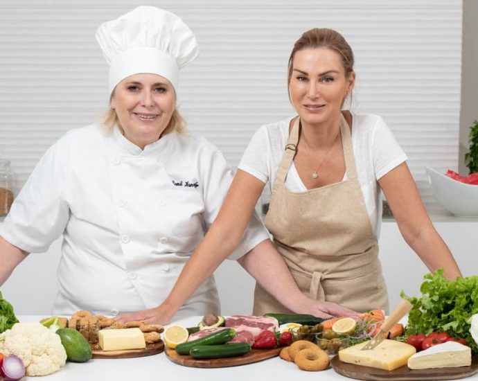 Two female chefs with variety of fresh ingredients in a kitchen setting, showcasing culinary preparation.